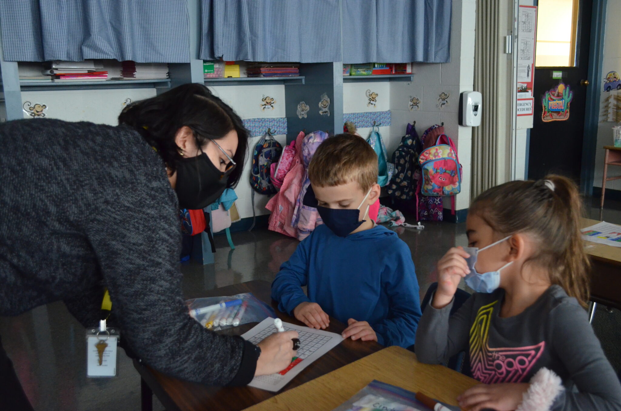 masked teacher helping two young masked students